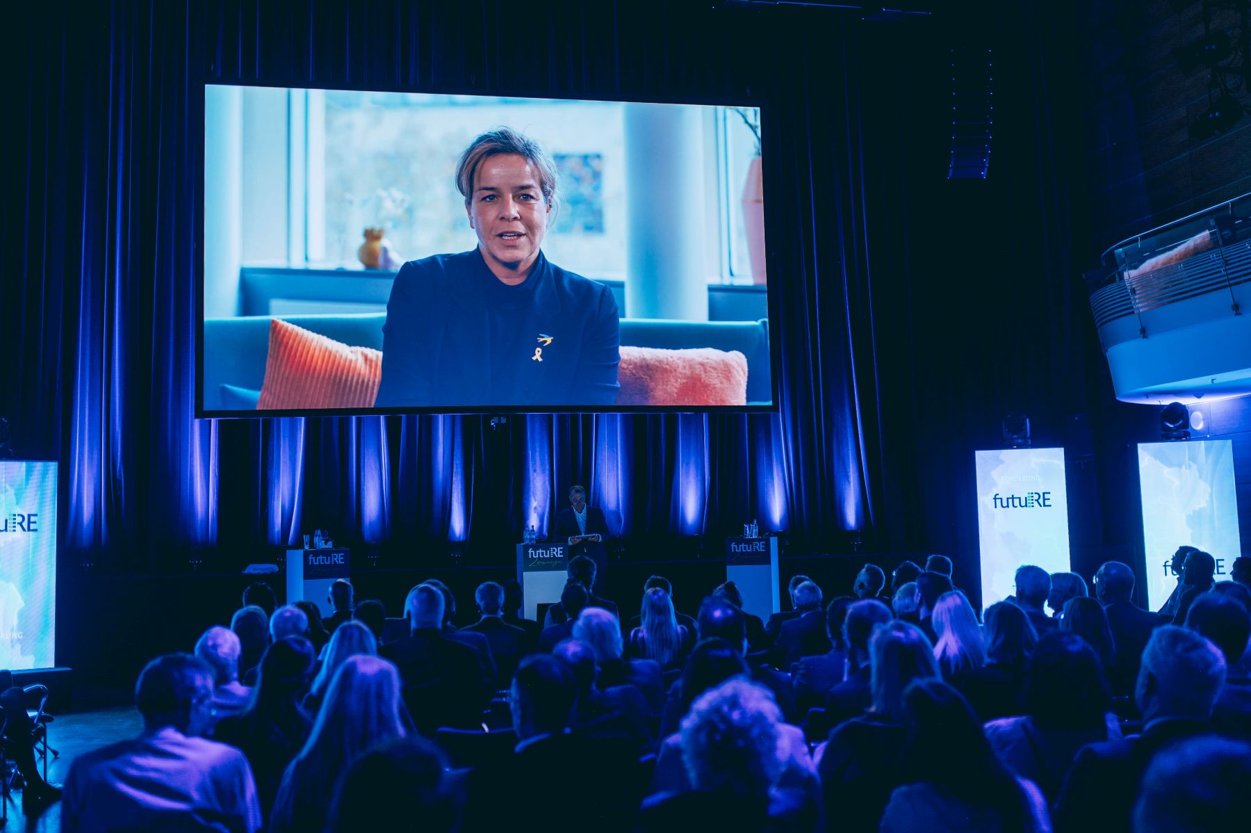 Bild von der Bühne, Blick auf die Leinwand. Schirmfrau des Wettbewerbs spricht zu den Teilnehmenden.